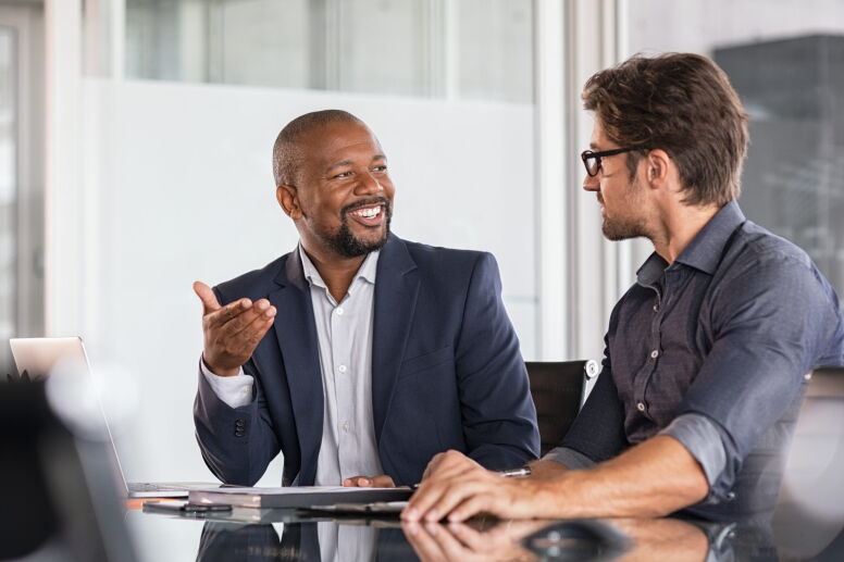 Two men in discussion at a table in an office setting. One man is wearing a navy blue blazer and smiling while gesturing; the other is wearing a dark blue shirt and glasses, listening attentively.
